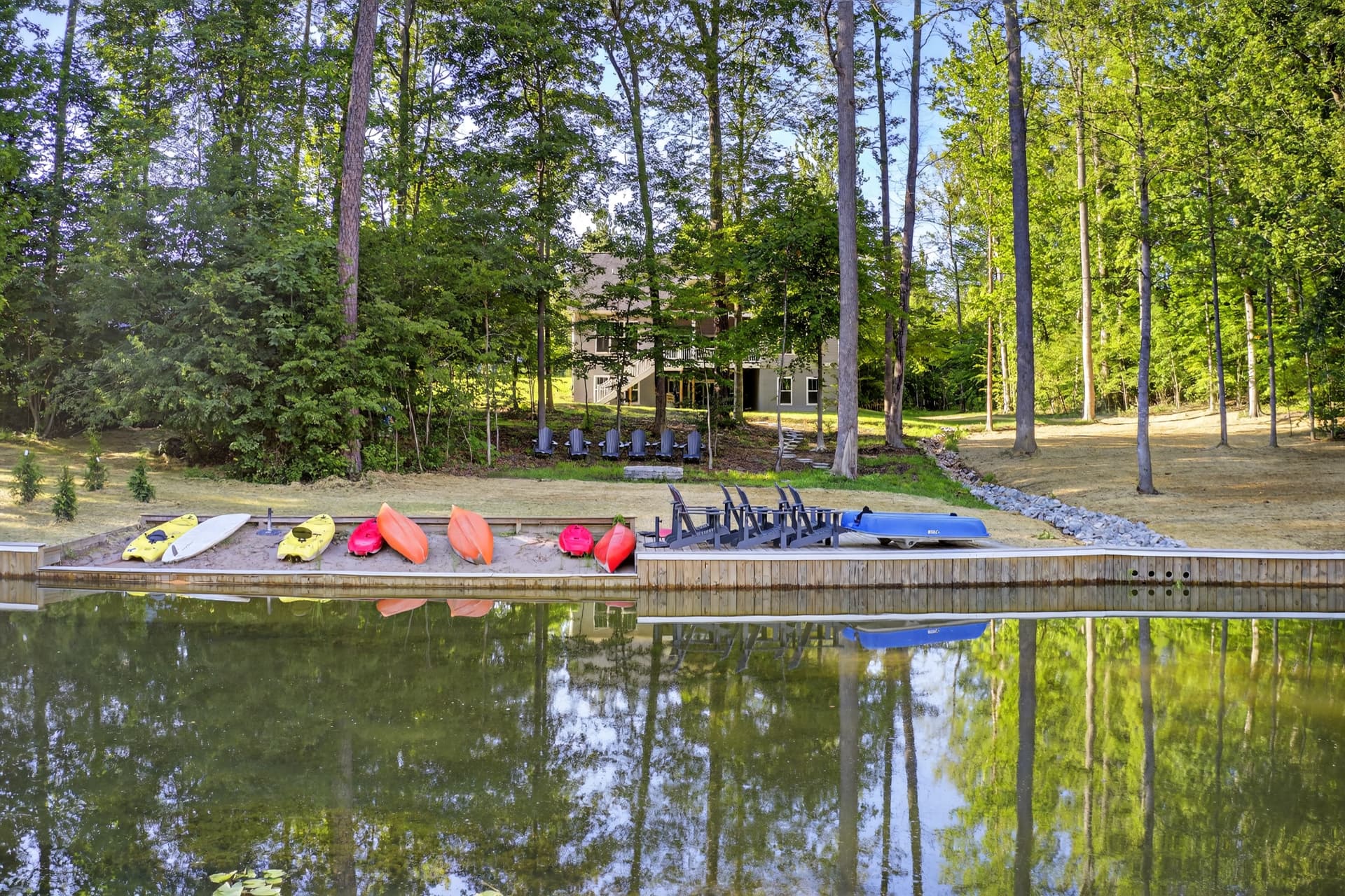 Kayaks and paddleboards on a lakeside beach beside a dock with seating and a wooded property behind it.
