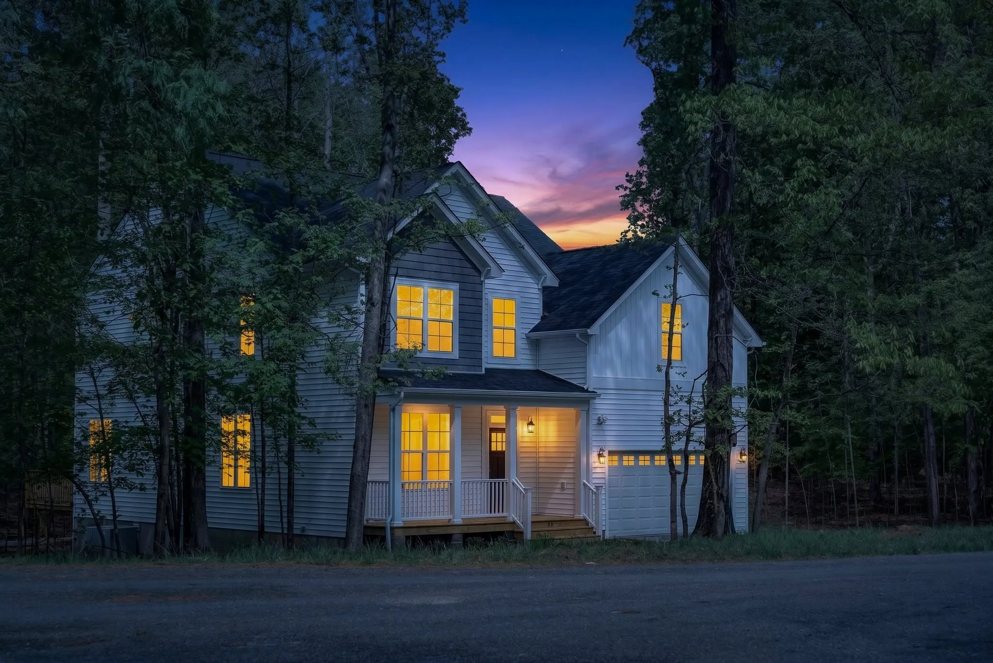 Virtual Twilight image of a two-story farmhouse surrounded by trees with every window glowing against a dusk sky