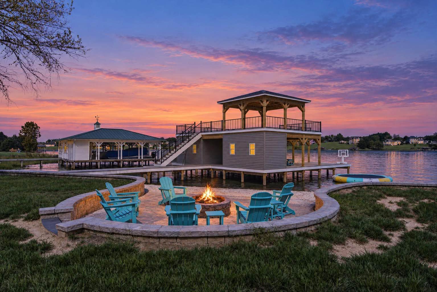 Blue chairs around a lit lakeside fire pit, with a boathouse and elevated pavilion beneath a colorful sunset sky.